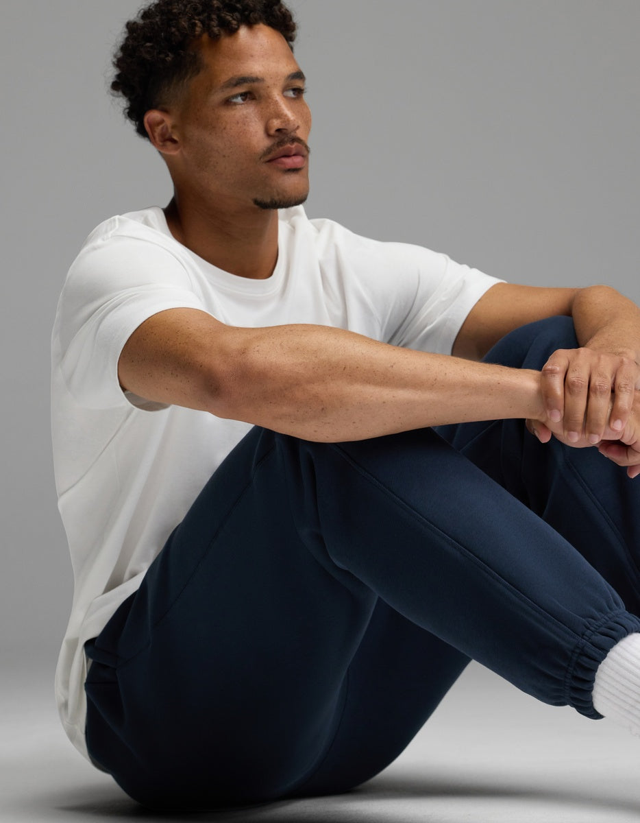 A man with short curly hair sits on the floor, arms resting on his knees, wearing a white t-shirt and Cozy Earth’s Men’s Cityscape Sweatpant in navy. He looks off to the side with a neutral expression against a plain gray background.