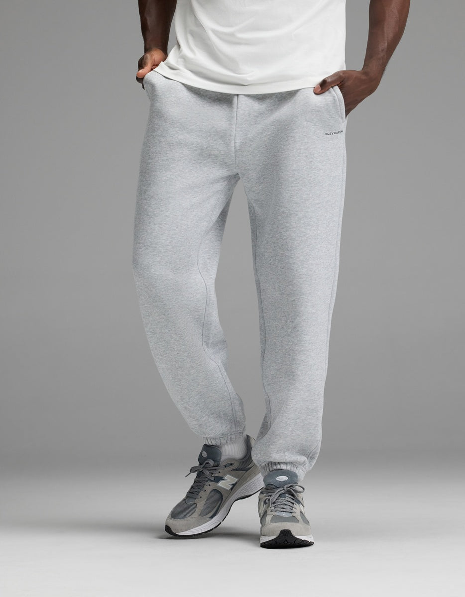 A person wearing a white shirt, Cozy Earth Men’s Cityscape Sweatpant in light gray, and gray sneakers stands with hands in pockets against a plain gray background.