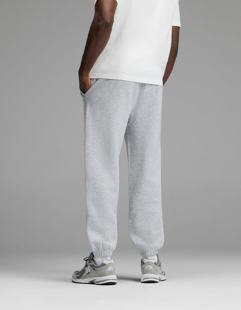 A person stands with hands in pockets, wearing a white T-shirt, Cozy Earth Men’s Cityscape Sweatpant in light gray, and gray sneakers. The background is plain gray. The image is shown from behind the knees up.