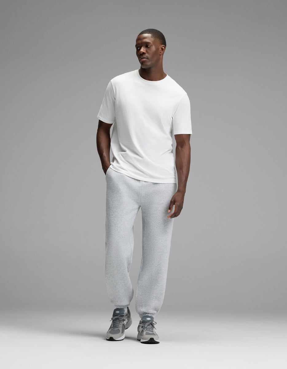A man stands against a plain gray background, wearing a white t-shirt, Cozy Earth Men’s Cityscape Sweatpant in light gray, and gray sneakers. His hands are in his pockets, and he is looking slightly to the side with a neutral expression.