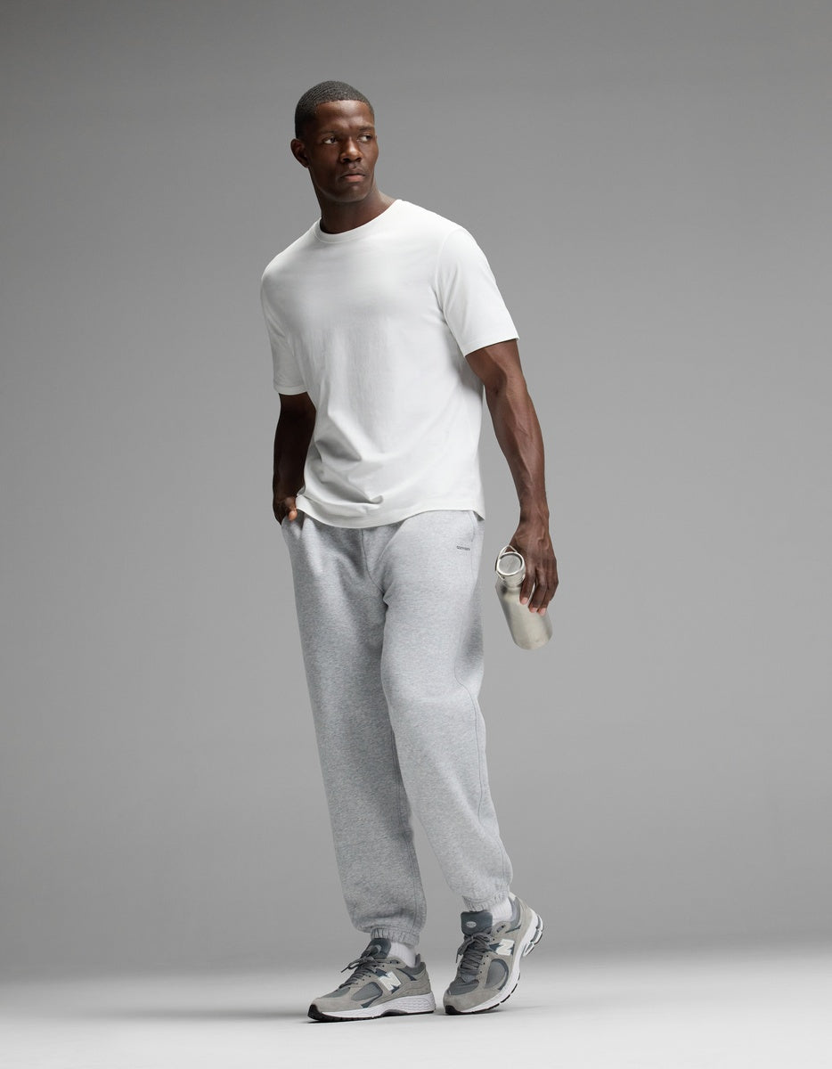 A man stands against a plain gray background, looking to the side, dressed in a white T-shirt and gray sneakers while holding a clear water bottle. He is wearing Cozy Earth Men’s Cityscape Sweatpant.