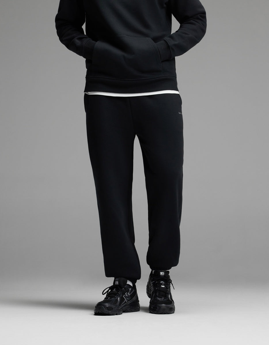 A person standing with hands in the pockets of Cozy Earth Men’s Cityscape Sweatpant in Jet Black, paired with a black sweatshirt, white shirt underneath, and black sneakers. The upper face is not visible.