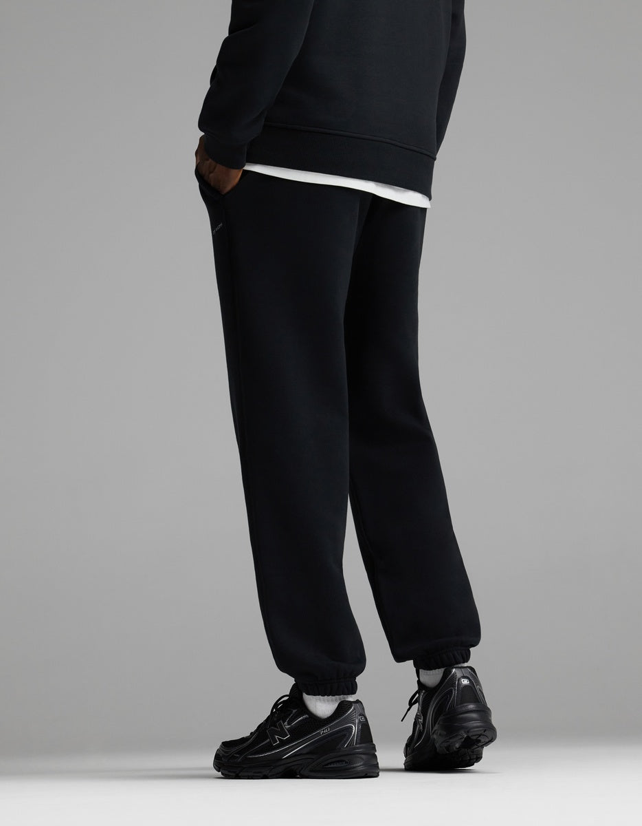 A person wearing Cozy Earth Men’s Cityscape Sweatpant in black stands facing away, with only their legs and lower torso visible, against a plain gray background.