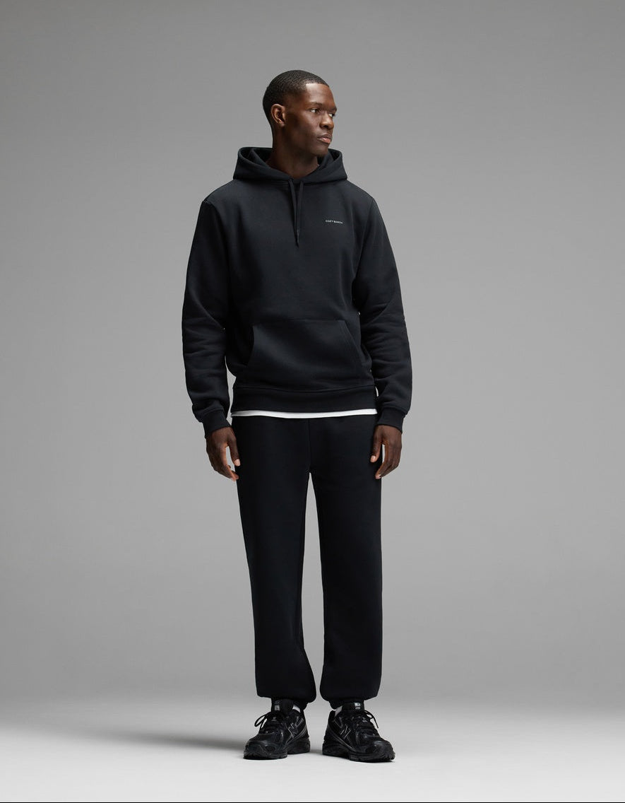 A man wearing Cozy Earth’s Men’s Cityscape Sweatpant in black, along with a matching black hoodie and sneakers, stands against a plain gray background, looking to his left.