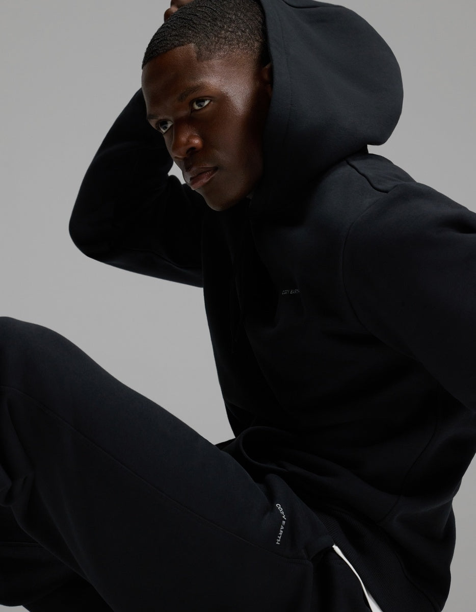 A man wearing Cozy Earth’s Men’s Cityscape Sweatpant in black sits in a relaxed pose against a plain gray background, looking to the side with one hand on his head.