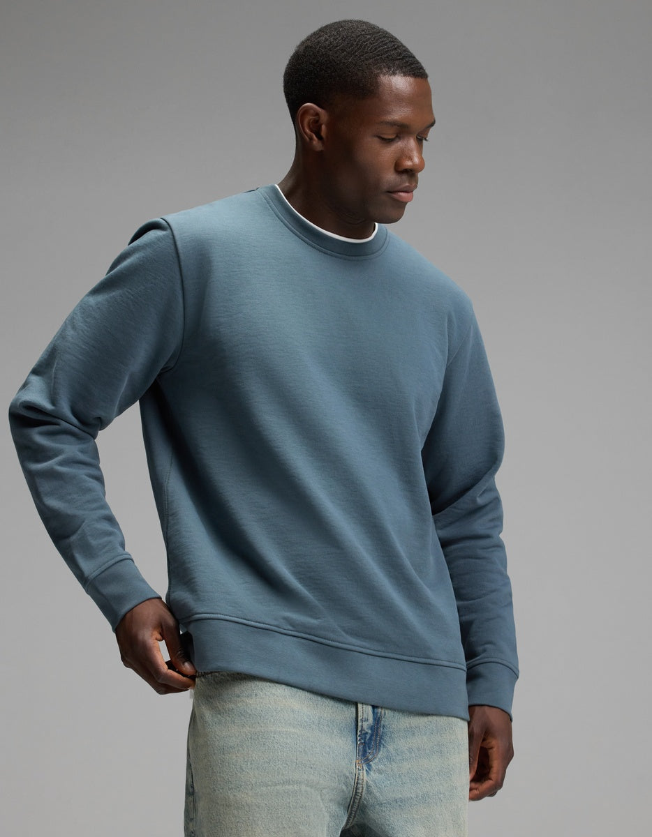A man wearing the Cozy Earth Men’s Dylan Crewneck and light blue jeans stands against a plain grey background, looking down as he adjusts his sweatshirt.|Color:Dawn