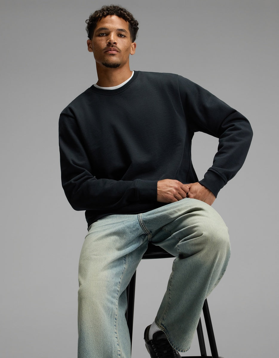 A man with short curly hair sits on a stool, wearing the Cozy Earth Men’s Dylan Crewneck, light blue jeans, and black sneakers, posing against a plain gray background.