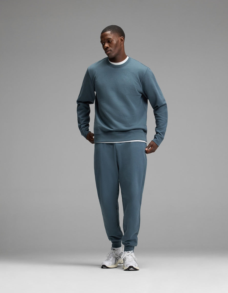 A man wearing the Cozy Earth Men’s Dylan Crewneck & Jogger Set in blue, paired with white sneakers, stands against a plain gray background, looking down with one hand in his pocket.|Color:Dawn