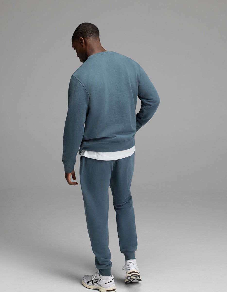 A person stands facing away against a plain gray background, wearing the Cozy Earth Men’s Dylan Crewneck & Jogger Set in blue-green over a white T-shirt and paired with white sneakers.