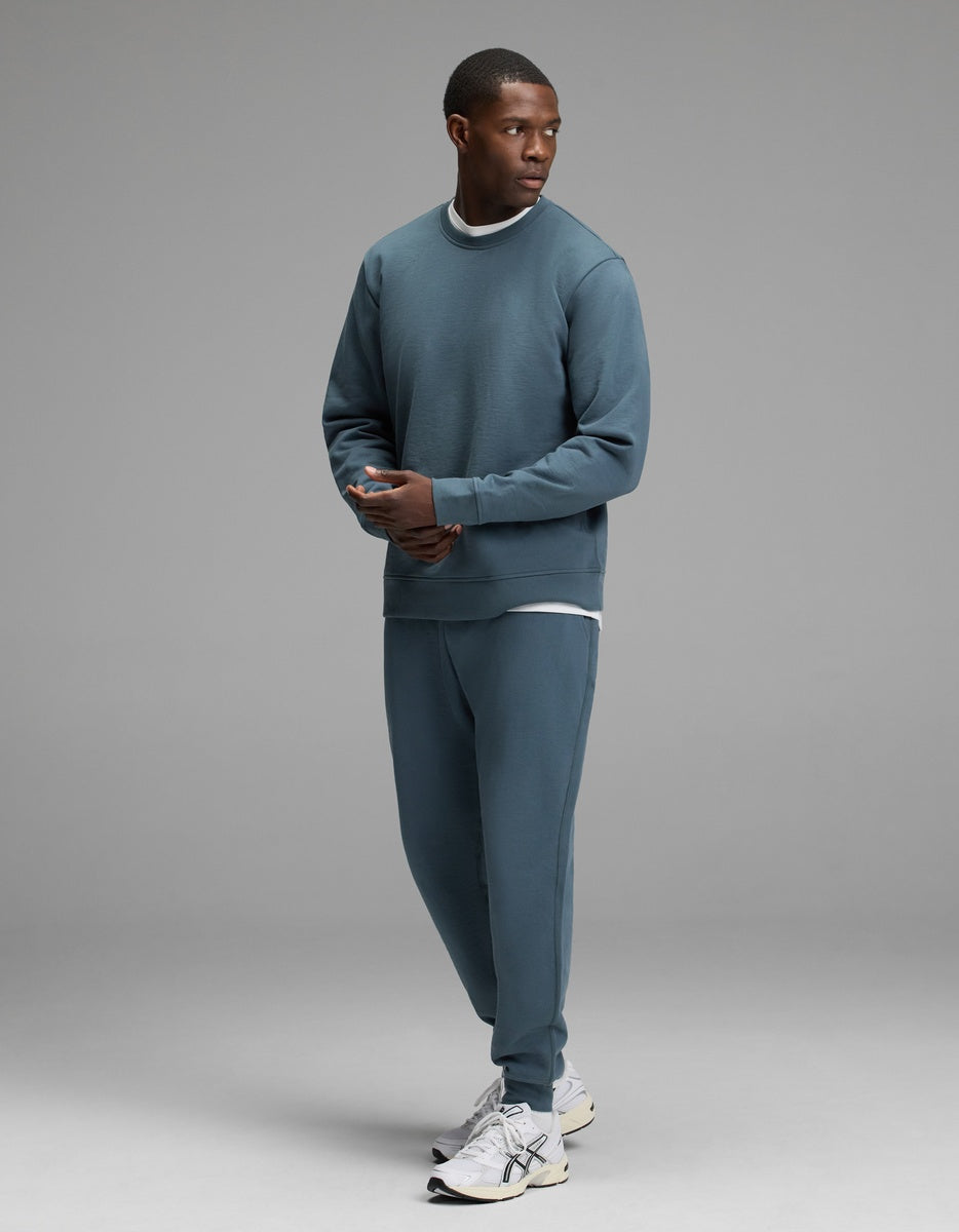 A man stands in a studio wearing the Cozy Earth Men’s Dylan Crewneck & Jogger Set paired with white sneakers. He looks to his left with his hands loosely clasped, against a plain light gray background.