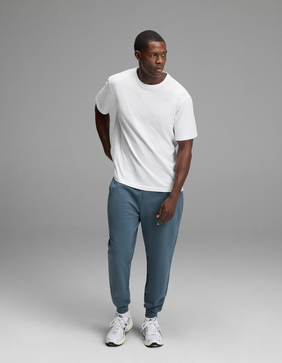 A man stands against a plain gray background, wearing a white t-shirt, Cozy Earth’s Men’s Dylan Jogger in blue, and white sneakers. He looks to his left with one hand behind his back and the other resting in front.