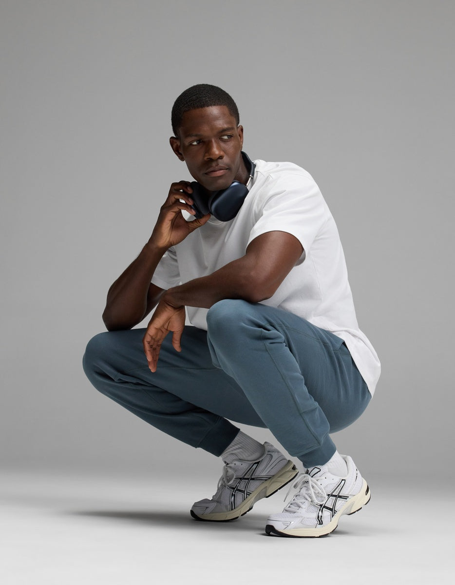 A man squats while wearing a white t-shirt, Cozy Earth Men’s Dylan Jogger in blue, white sneakers, and black headphones around his neck, looking to the side against a plain gray background.