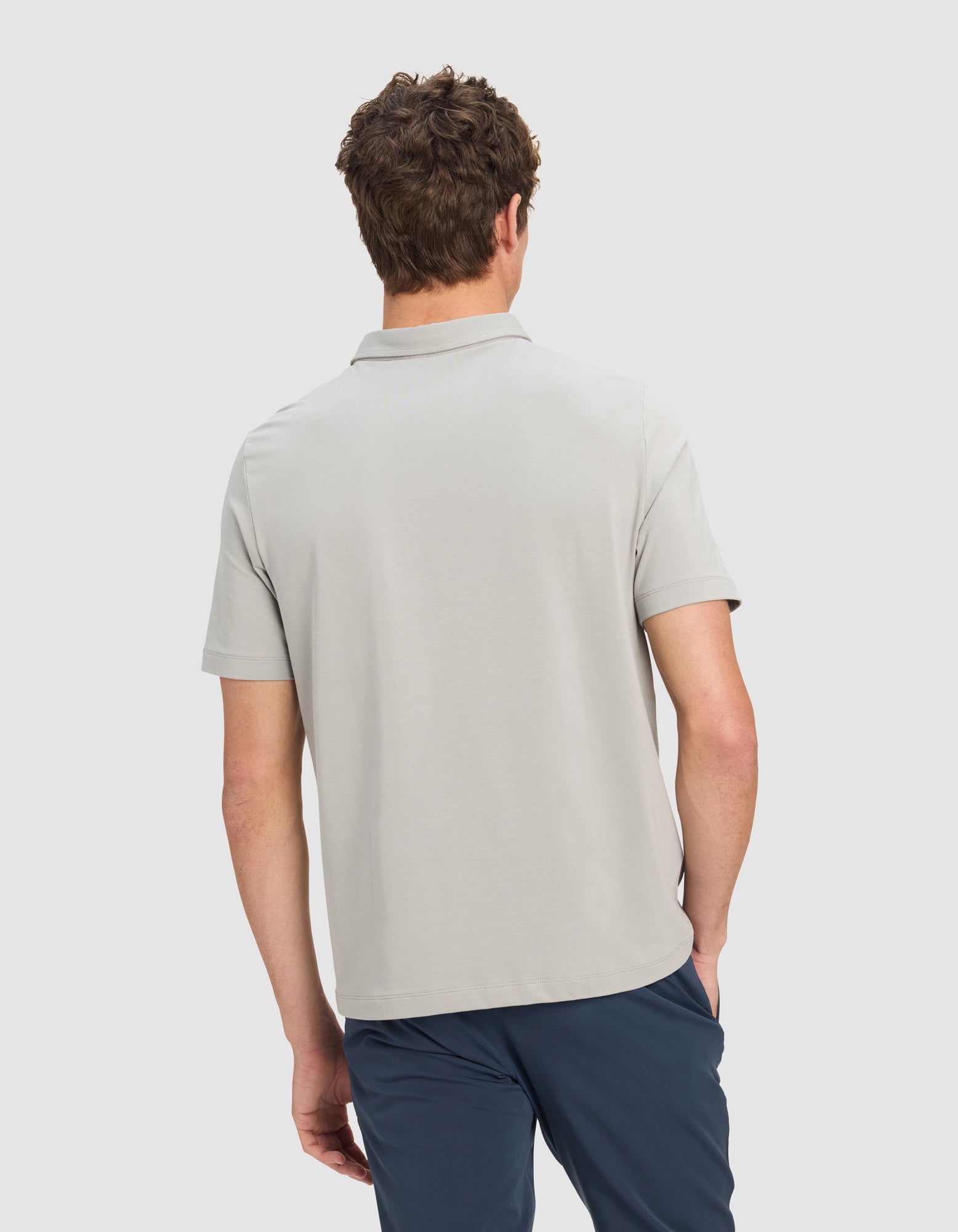 A man with short curly hair is shown from the back wearing the Cozy Earth Men's Everyday Polo in light gray and dark blue pants, standing against a plain light background.