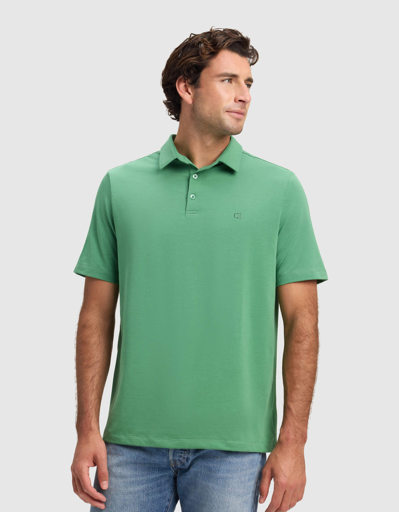 A man models the Cozy Earth Men's Everyday Polo in green with blue jeans, standing against a white background and gazing to his left with a relaxed expression.