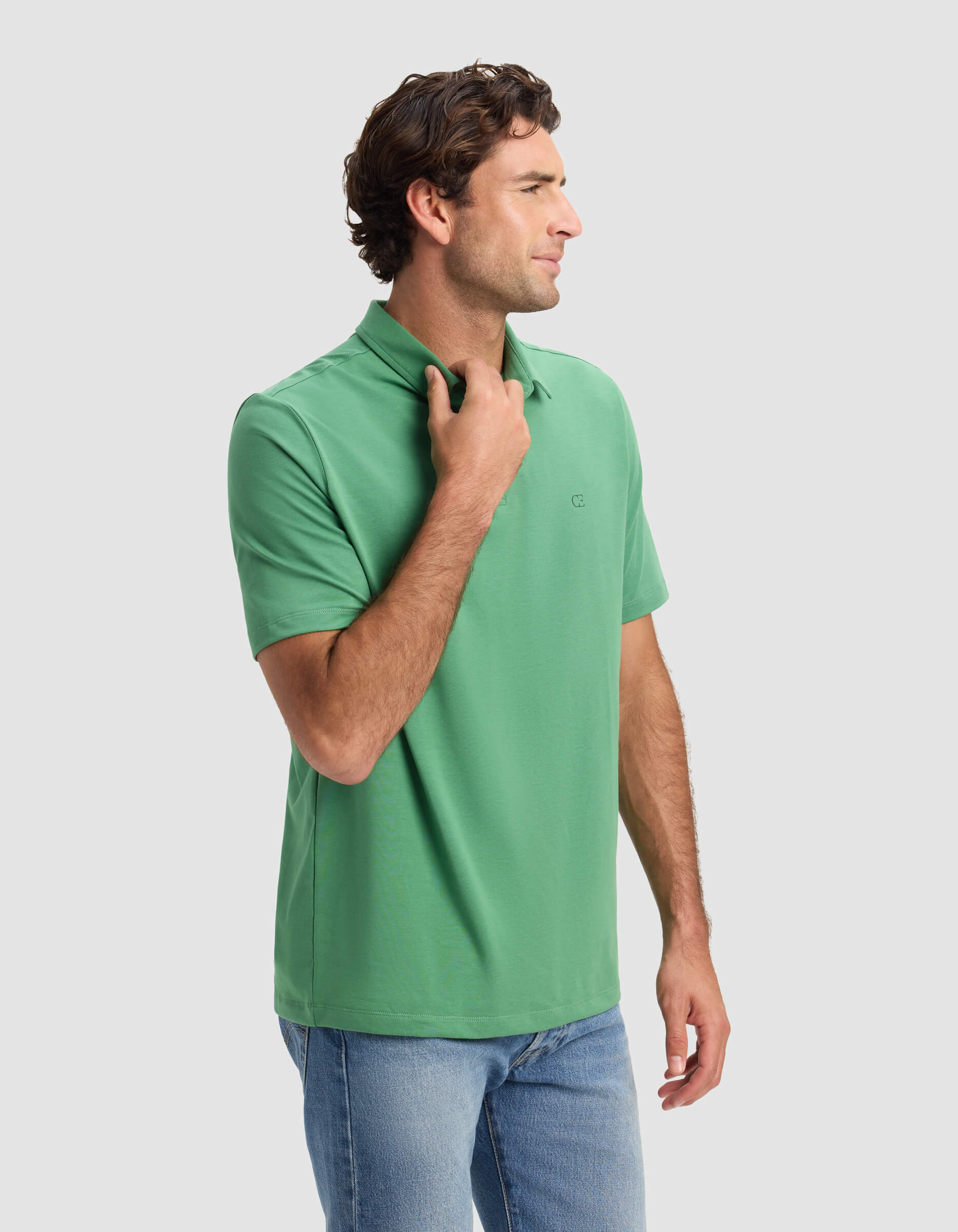 A man with wavy brown hair wears the Cozy Earth Men's Everyday Polo in green and blue jeans, standing sideways with one hand on the collar against a plain light gray background.|Color:Vine