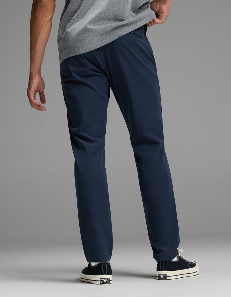 A man stands facing away against a plain background, wearing the Cozy Earth Men’s Everywhere Pant in navy blue, paired with a grey shirt and black sneakers with white soles, one hand resting on his hip.