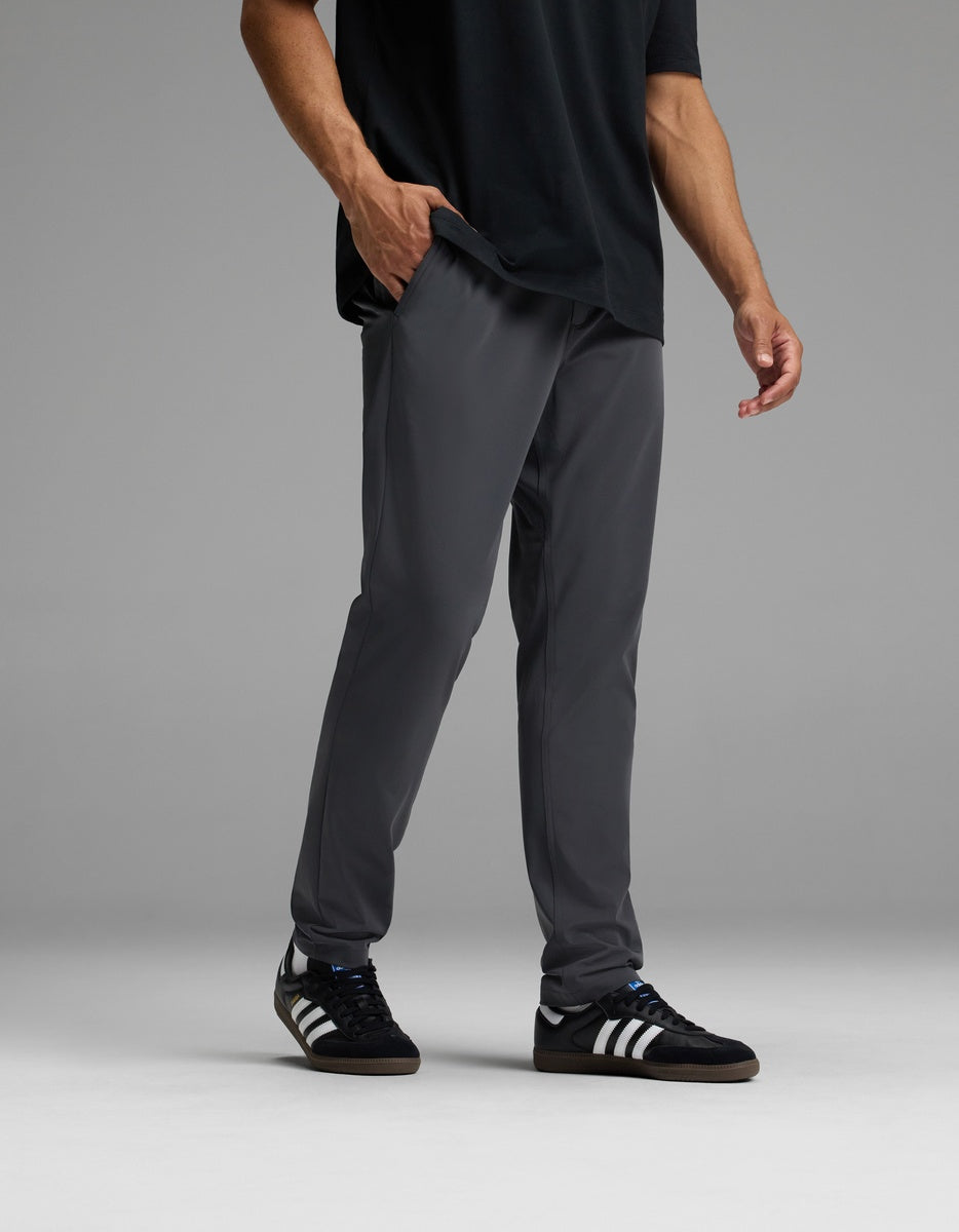 Wearing Cozy Earth’s Men’s Everywhere Pant in dark gray, along with a black T-shirt and black Adidas sneakers with white stripes, the individual stands against a plain gray background. Their right hand rests in their pocket, and their head is out of frame.|Color:Graphite