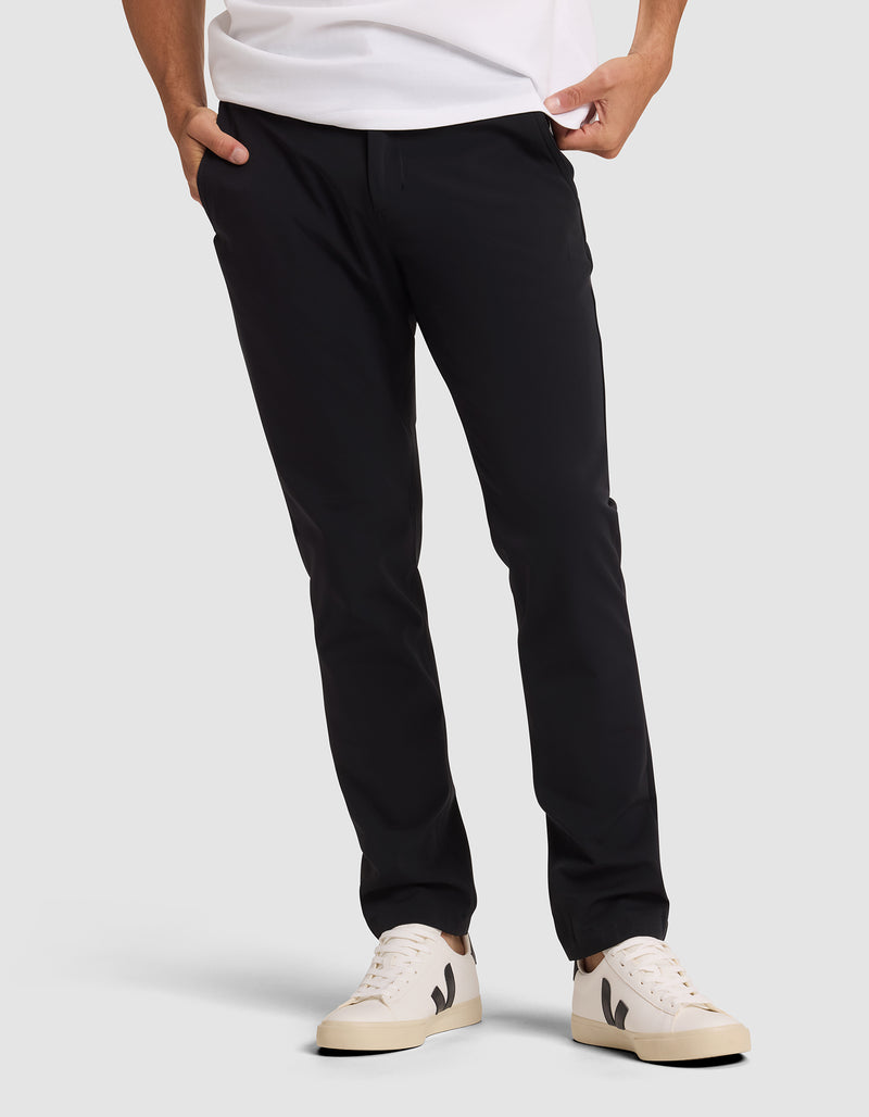 A person wearing Cozy Earth's Men's Everywhere Pant in black, a white t-shirt, and cream-colored sneakers with black detailing stands with hands in pockets against a plain light background.