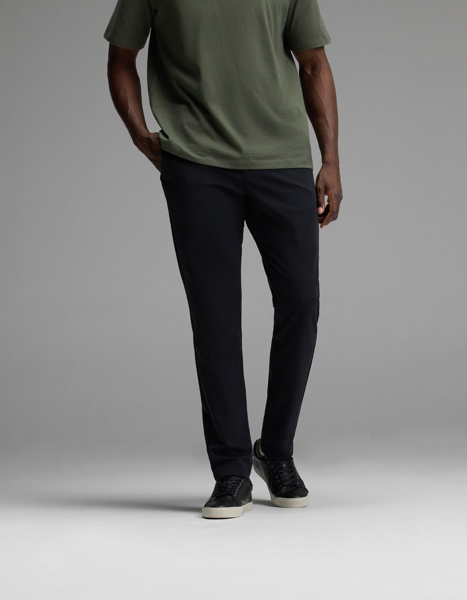 A person wearing Cozy Earth’s Men’s Everywhere Pant in black, paired with a short-sleeve olive green shirt and black sneakers with white soles, stands against a plain gray background. Their face is not visible.