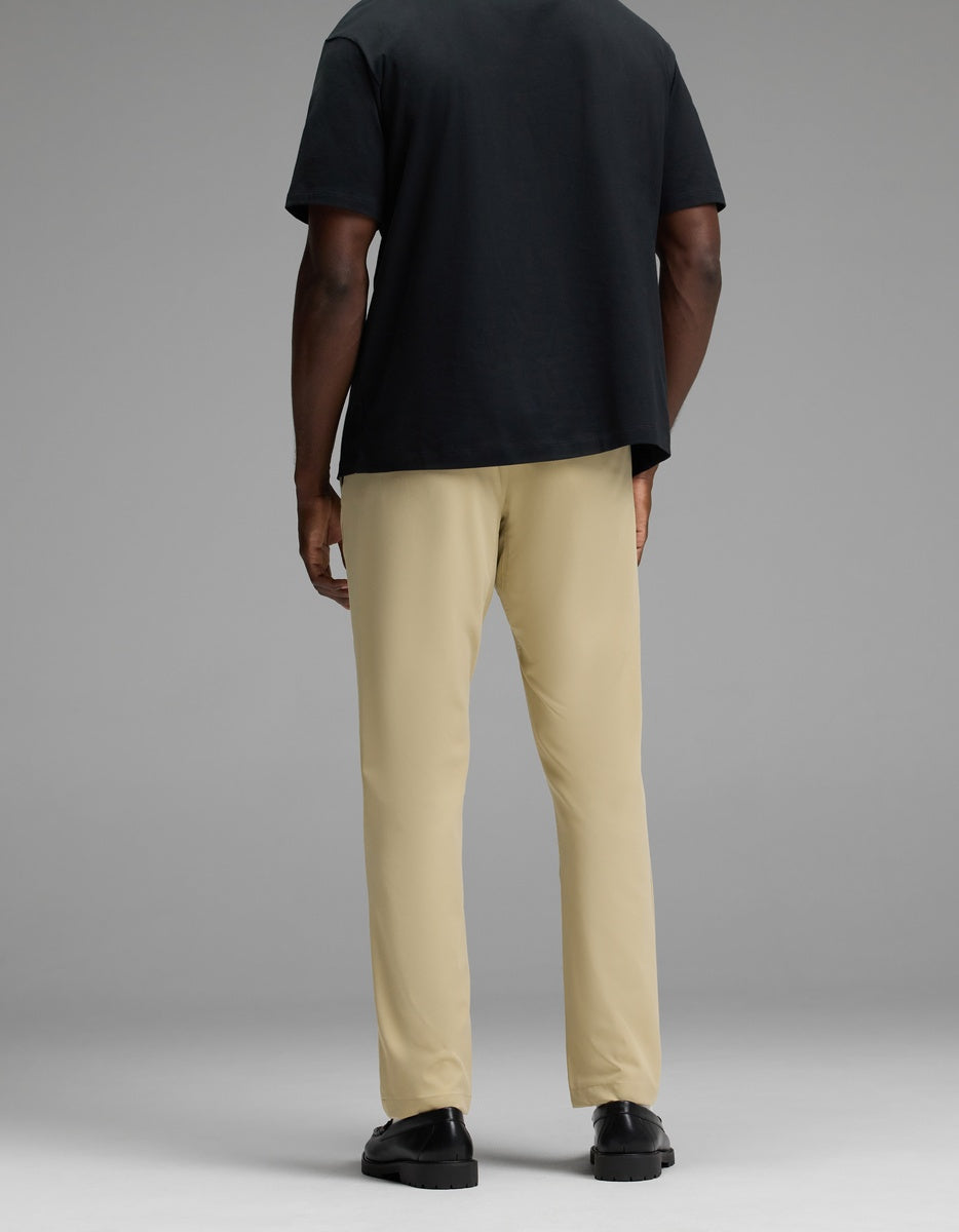 A person stands facing away, wearing Cozy Earth’s Men’s Everywhere Pant in beige, a black t-shirt, and black shoes against a plain gray background.