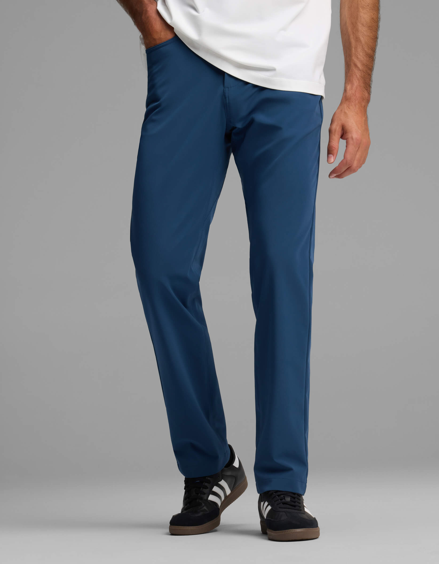 A person wearing Cozy Earth Men's Everywhere Pant - Relaxed Fit in blue, paired with a white shirt and black sneakers with white stripes, stands against a plain gray background.|Color:Obsidian