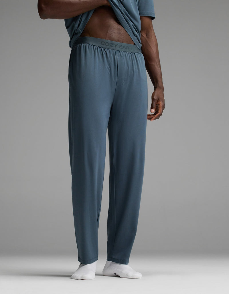 A person is wearing Cozy Earth Men’s First Layer Pajama Pants in blue, featuring a waistband with “COZY EARTH” branding. They are lifting their shirt to reveal the branded waistband and are also wearing white socks. The background is plain gray.