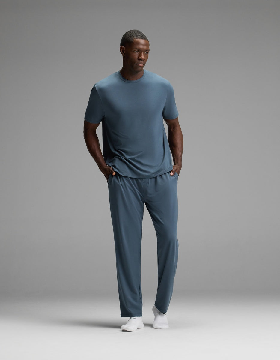 A man stands with his hands in his pockets, wearing the Cozy Earth Men’s First Layer Pajama Pant in blue paired with a matching t-shirt and white shoes, set against a plain gray background.