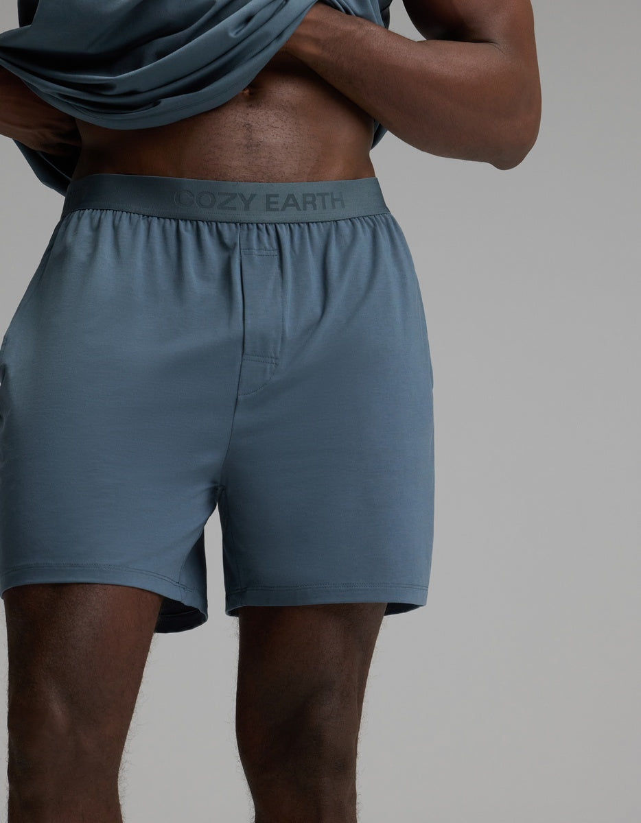 A person wears Cozy Earth’s Men’s First Layer Pajama Short in Dawn, featuring “Cozy Earth” on the waistband, lifting their shirt to reveal their torso and legs against a plain background.