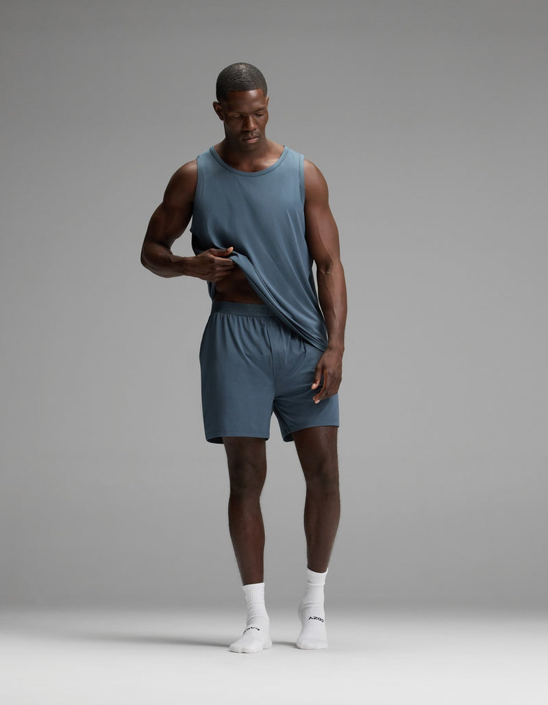 A man wearing Cozy Earth’s Men’s First Layer Pajama Short in blue stands on a gray background, paired with a matching sleeveless blue shirt and white socks, gently lifting his shirt while looking down.