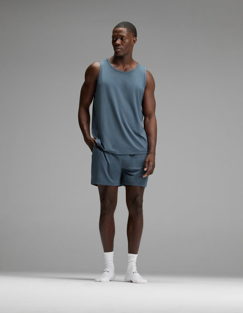 A man wearing the Cozy Earth Men’s First Layer Pajama Tank in blue, with matching shorts and white socks, stands against a plain gray background, looking to his left with one hand in his pocket.