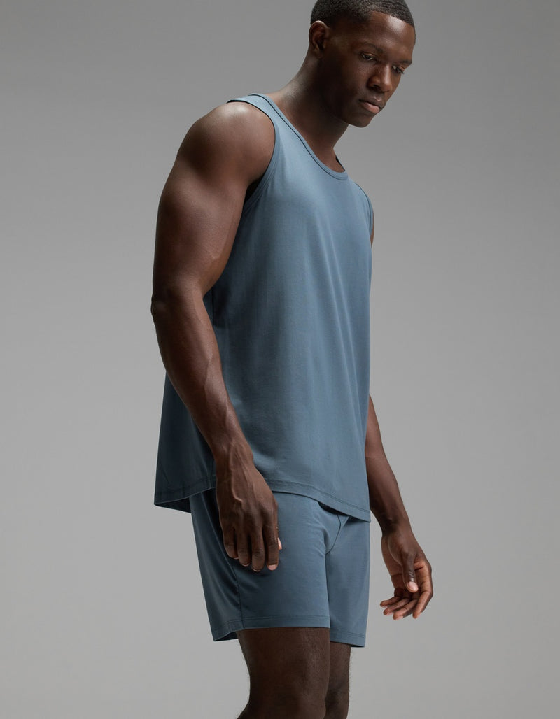 A man stands in a studio wearing the Cozy Earth Men’s First Layer Pajama Tank and matching shorts in blue, looking downward with a neutral expression against a plain gray background.