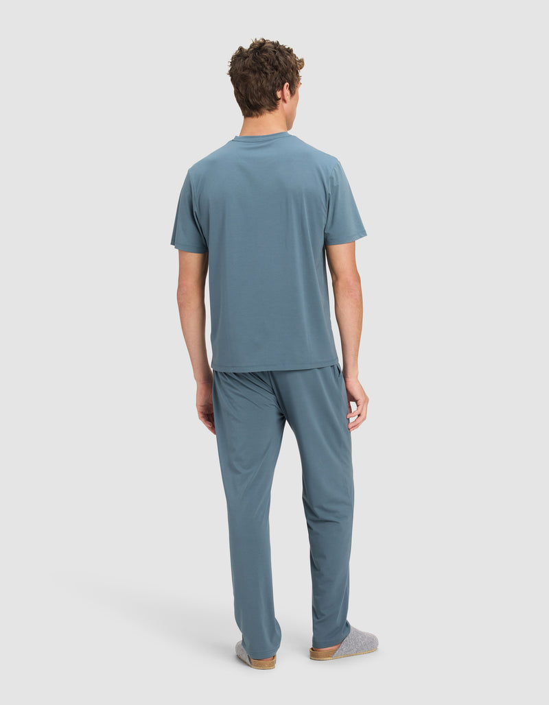 A man faces away, wearing the Cozy Earth Men's First Layer Pajama Short Sleeve Tee & Pant Set in blue with gray slippers, against a plain white background.