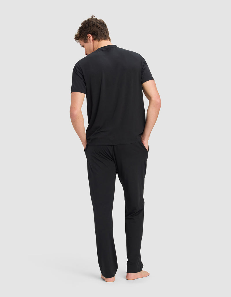 A man stands barefoot with his hands in his pockets, wearing the Cozy Earth Men's First Layer Pajama Short Sleeve Tee & Pant Set in black, facing away against a plain light gray background. His head is slightly tilted downward.