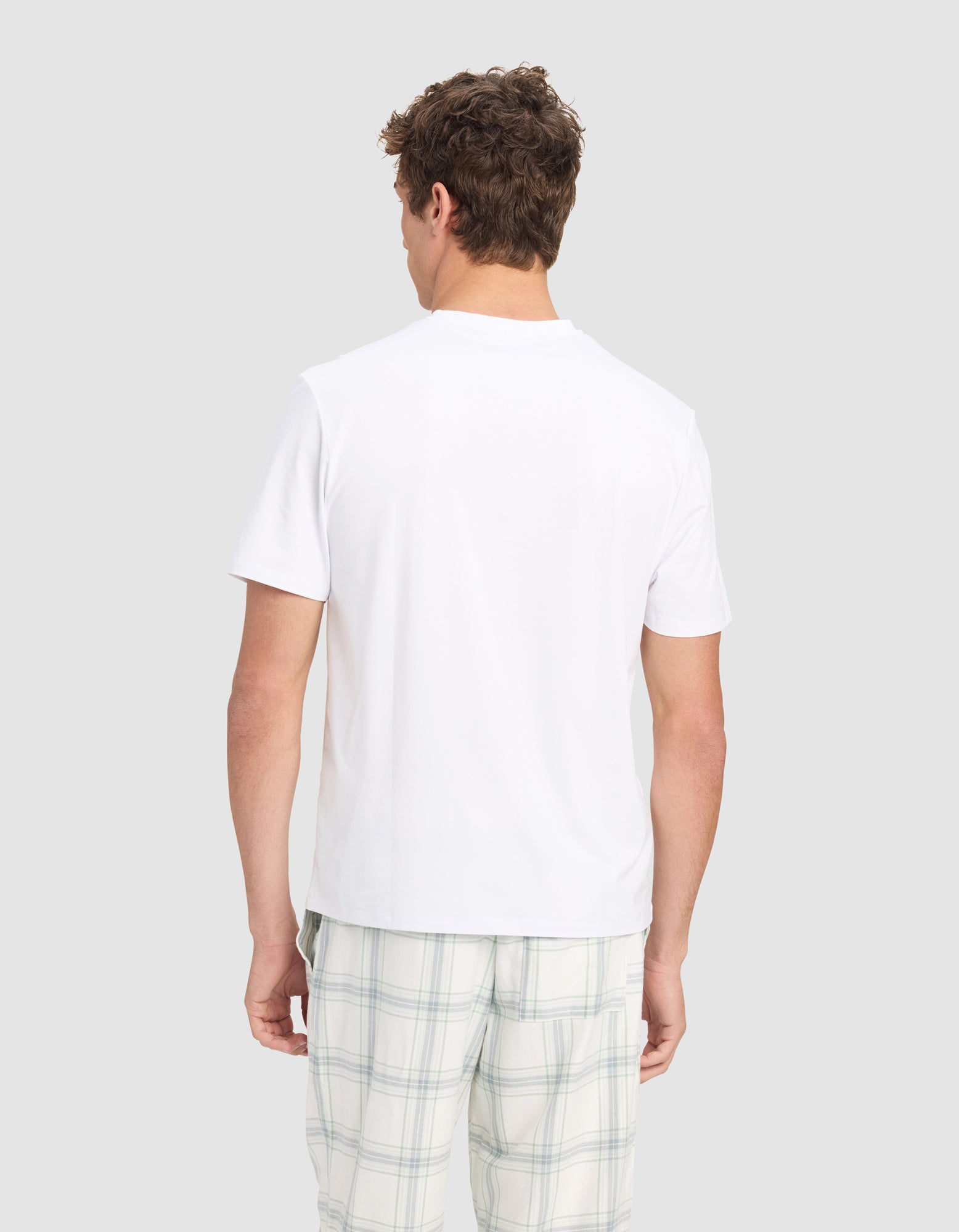 A man faces away from the camera, wearing the Cozy Earth Men's First Layer Pajama Short Sleeve Tee and light plaid shorts, set against a plain light gray background.
