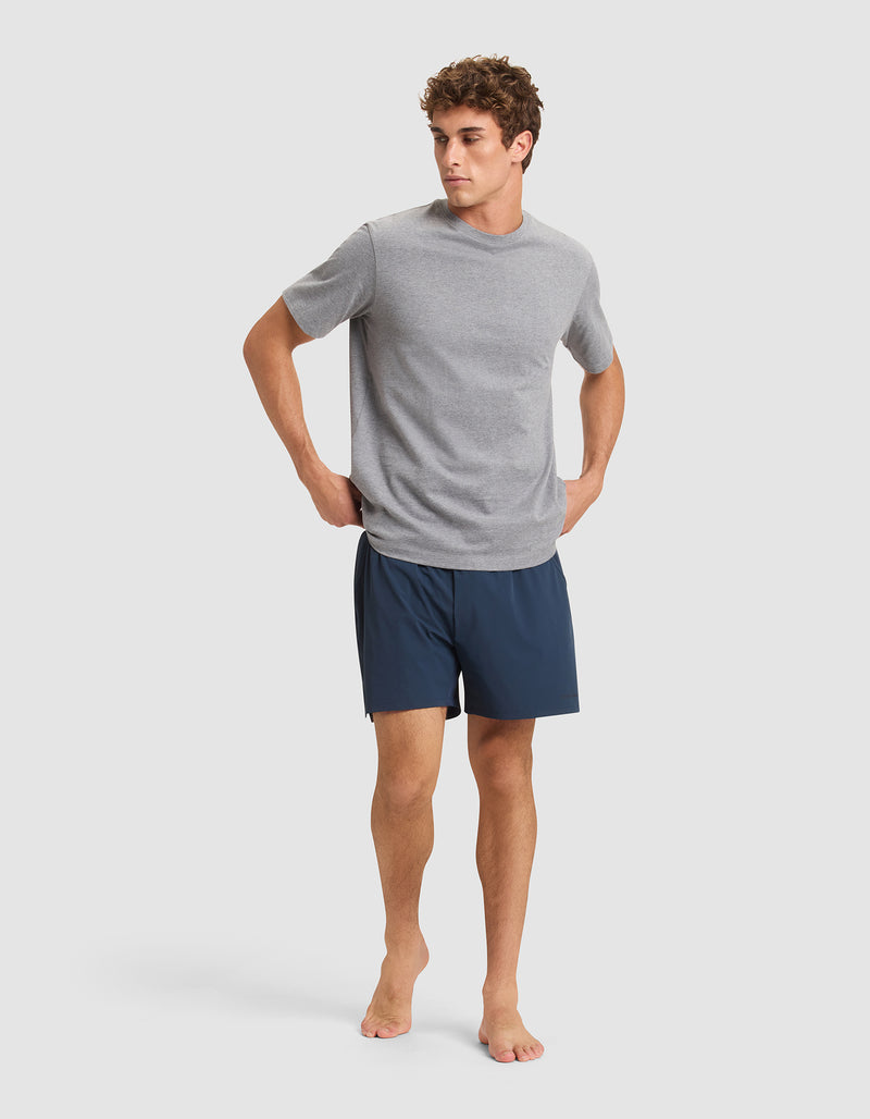 A man stands barefoot against a plain background, wearing a light gray short-sleeve T-shirt and Cozy Earth Men's Day & Night Shorts 5" in dark blue, with his hands resting on his hips.