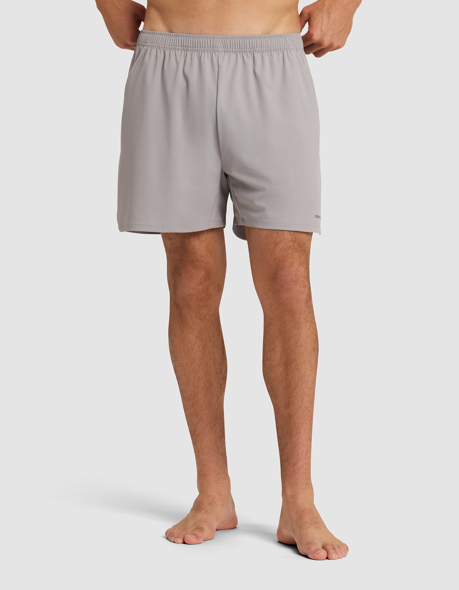 A person stands barefoot wearing the Cozy Earth Men's Day & Night Short 5" in light gray, featuring an elastic waistband. The image is cropped from the waist down against a plain white background. 