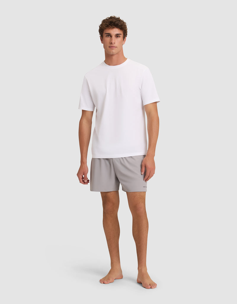 A young man stands barefoot against a light grey background, facing forward with a neutral expression. He is wearing Cozy Earth Men's Day & Night Shorts 5" in light grey and a plain white short-sleeve t-shirt.