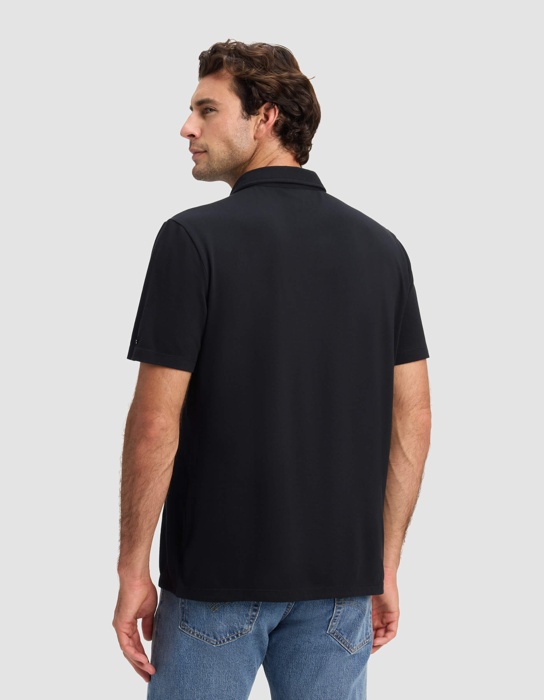 A man faces away wearing the Cozy Earth Men's Pique Polo in black with blue jeans against a light gray background, looking over his left shoulder with a neutral expression.