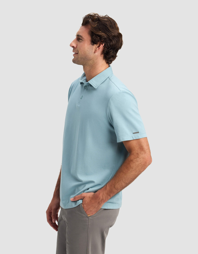 A man models the Cozy Earth Men's Pique Polo in light blue, paired with gray pants. He stands in profile with one hand in his pocket, facing left against a plain light gray background.