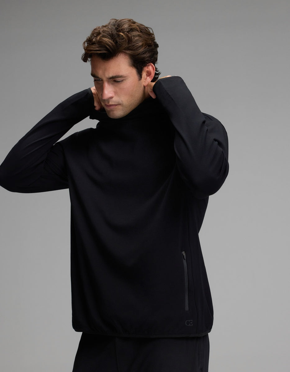 A man with wavy brown hair wears the Cozy Earth Men’s Recovery Hoodie in black, lifting its collar over his head. The background is plain gray, and the hoodie features a zippered pocket on the side.