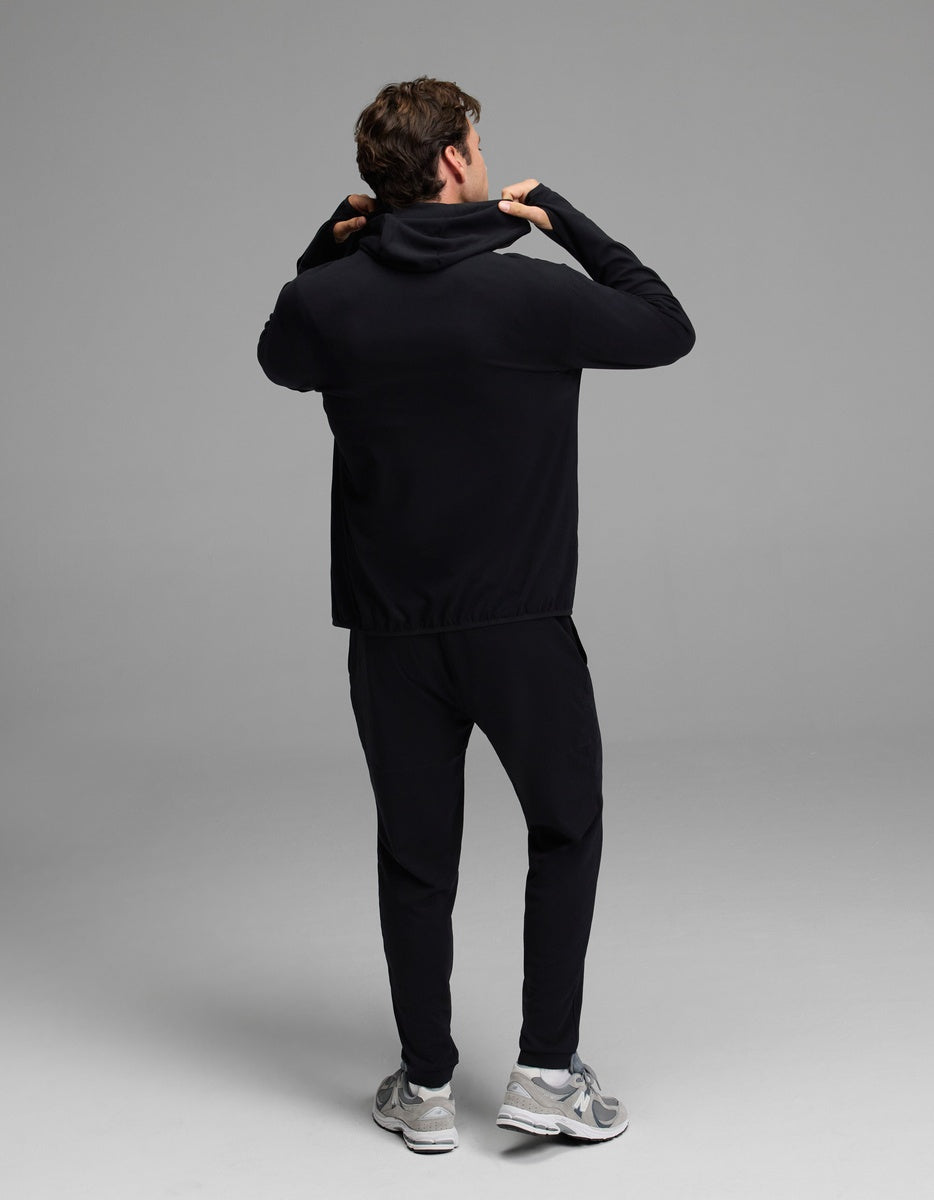 A person faces away while adjusting the hood of a black Men’s Recovery Hoodie & Jogger Set from Cozy Earth, paired with gray sneakers, posing in a simple, well-lit studio against a plain gray background.