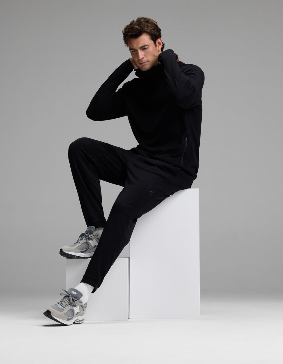 A man wearing Cozy Earth’s Men’s Recovery Jogger and gray sneakers sits on white blocks with one knee raised, hands behind his neck, looking downward against a plain gray background.