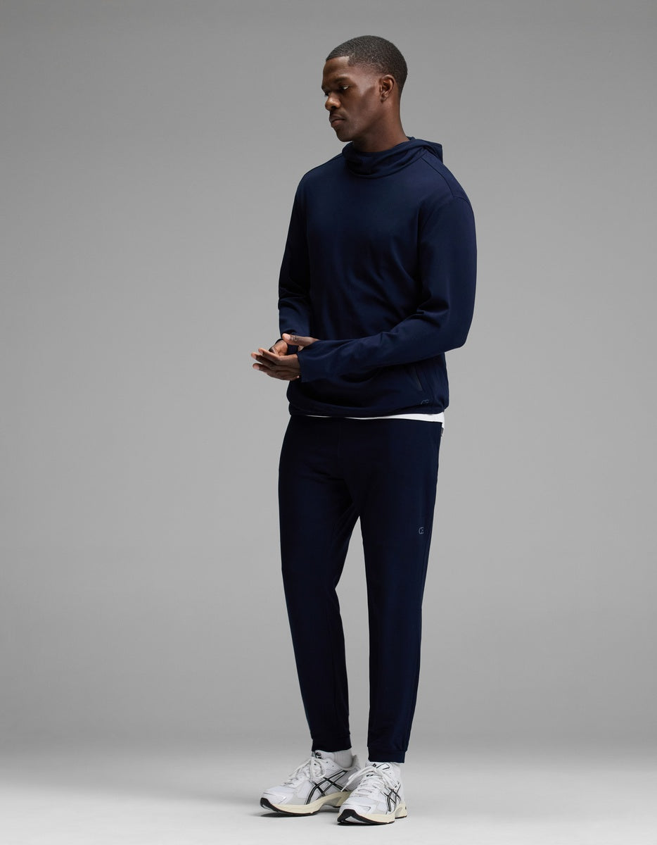 A man stands wearing the Cozy Earth Men’s Recovery Jogger in navy, paired with a matching navy hooded sweatshirt and white athletic sneakers, set against a plain gray background.