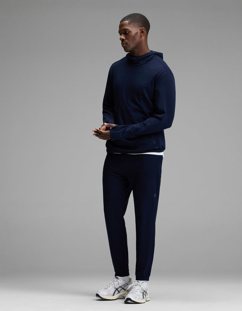 A man stands wearing the Cozy Earth Men’s Recovery Jogger in navy, paired with a matching navy hooded sweatshirt and white athletic sneakers, set against a plain gray background.