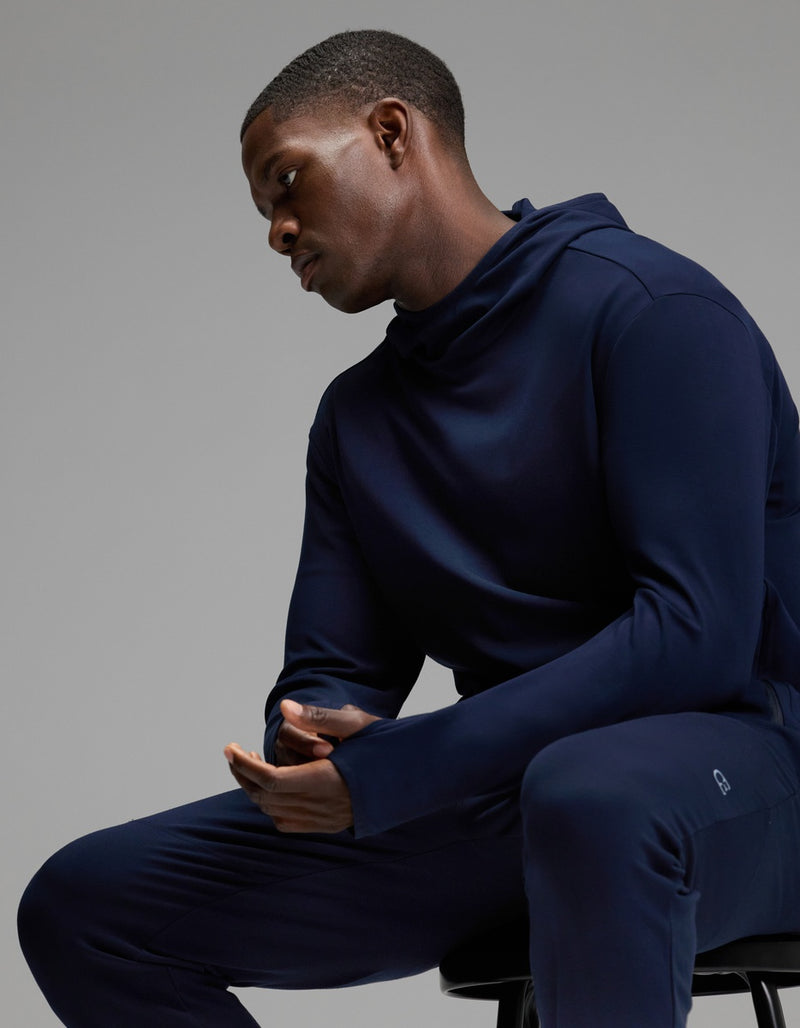 A man wearing Cozy Earth’s Men’s Recovery Jogger in dark blue sits on a stool with his hands clasped, looking down thoughtfully against a plain gray background.