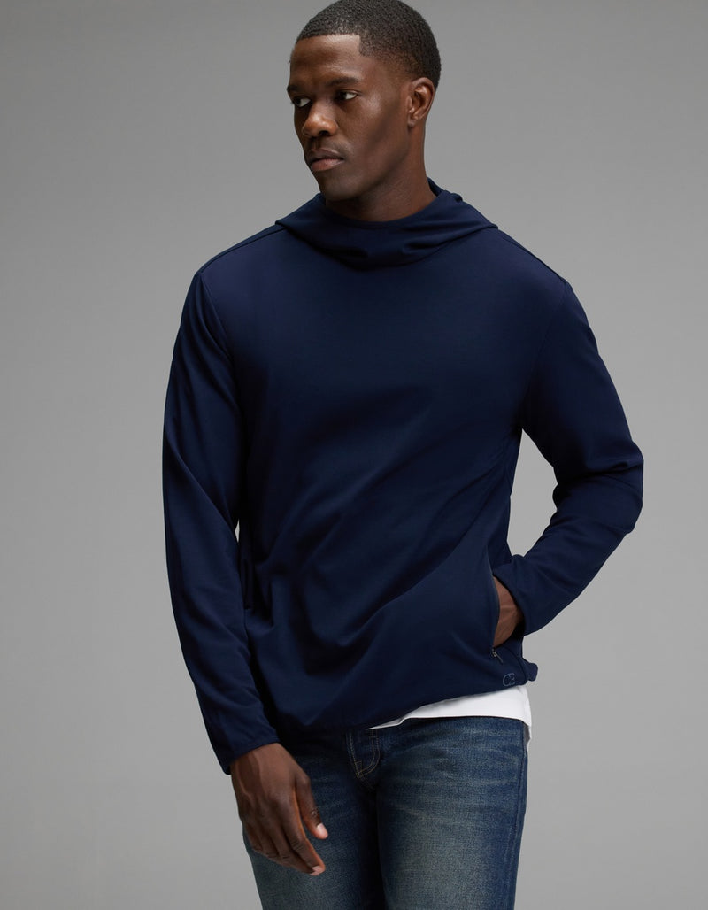 A person wearing the Cozy Earth Men’s Recovery Hoodie in navy stands against a plain gray background, with one hand in their pocket and looking slightly to the side.