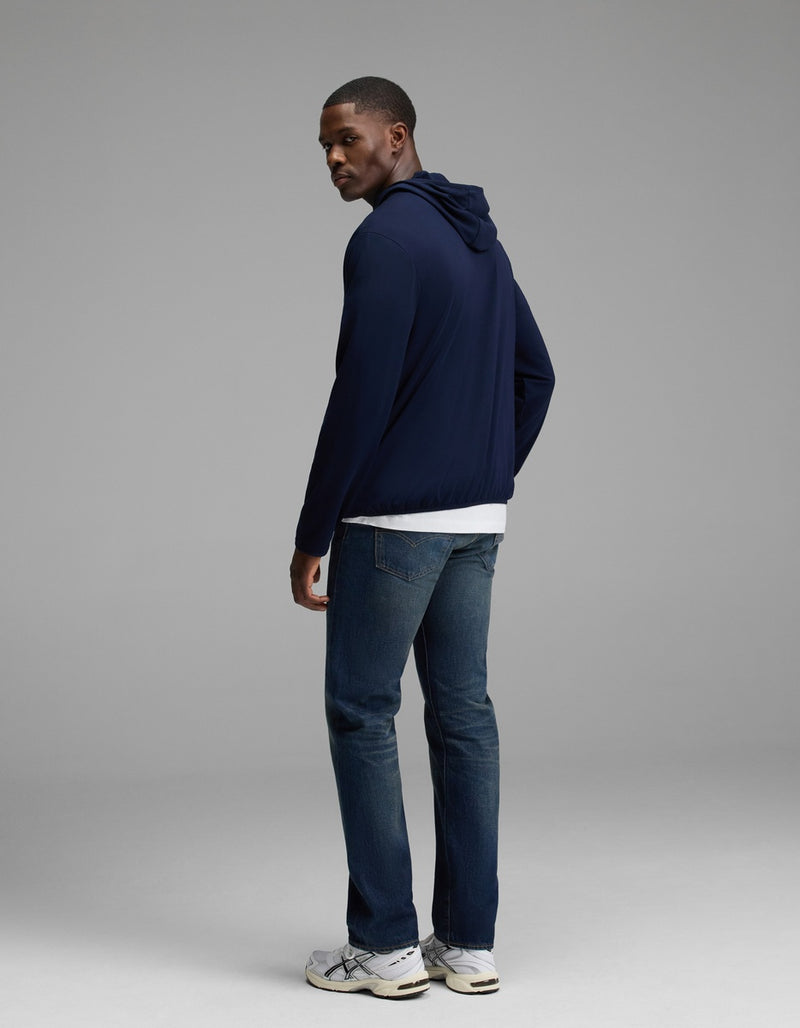 A man stands in a studio, facing slightly away, wearing the Cozy Earth Men’s Recovery Hoodie in navy over a white shirt, paired with blue jeans and white sneakers against a plain gray background.