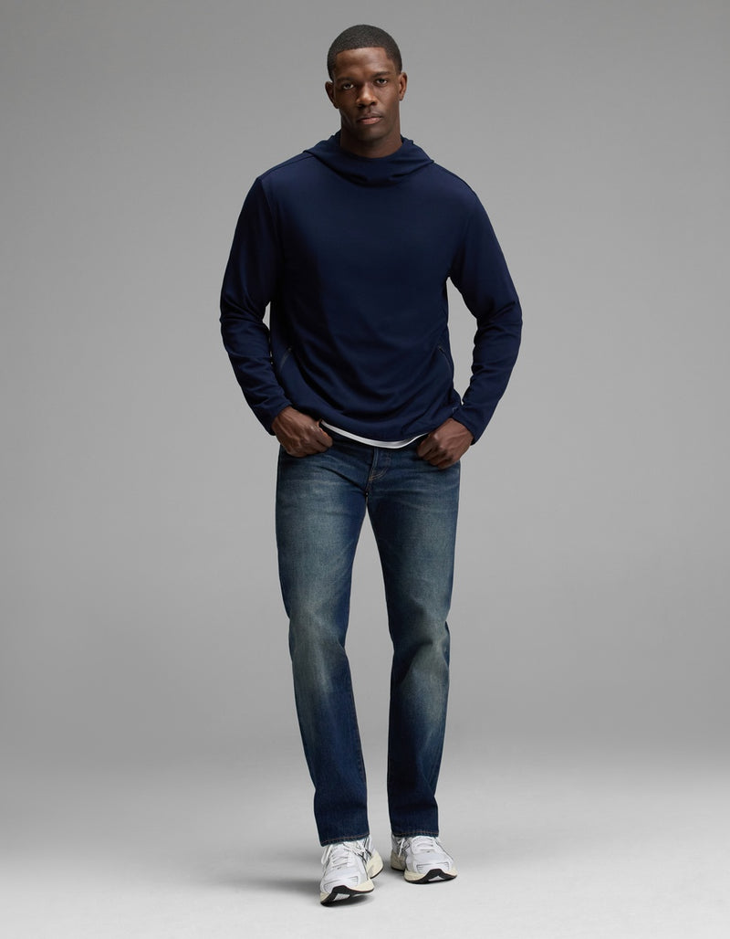 A man stands facing forward against a plain grey background, wearing the Cozy Earth Men’s Recovery Hoodie in navy, blue jeans, and white sneakers.