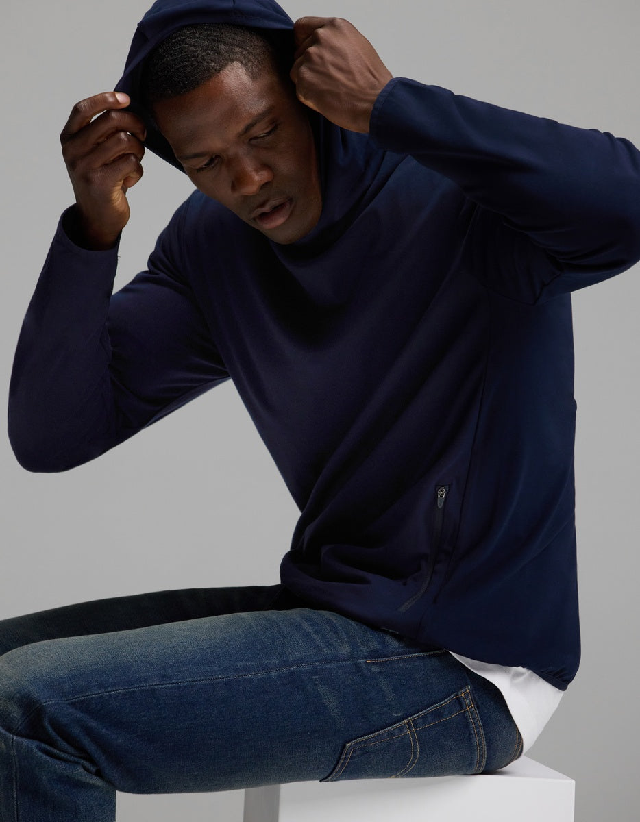 A person wearing the Cozy Earth Men’s Recovery Hoodie and blue jeans sits on a white block, pulling the hood over their head, against a plain gray background.|Color:Navy