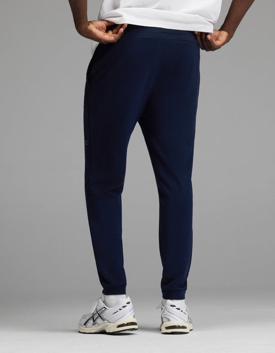 A man models the Cozy Earth Men’s Recovery Jogger in navy, paired with white sneakers featuring black accents and a white shirt, standing against a plain gray backdrop. The photo highlights the back view of the lower body.